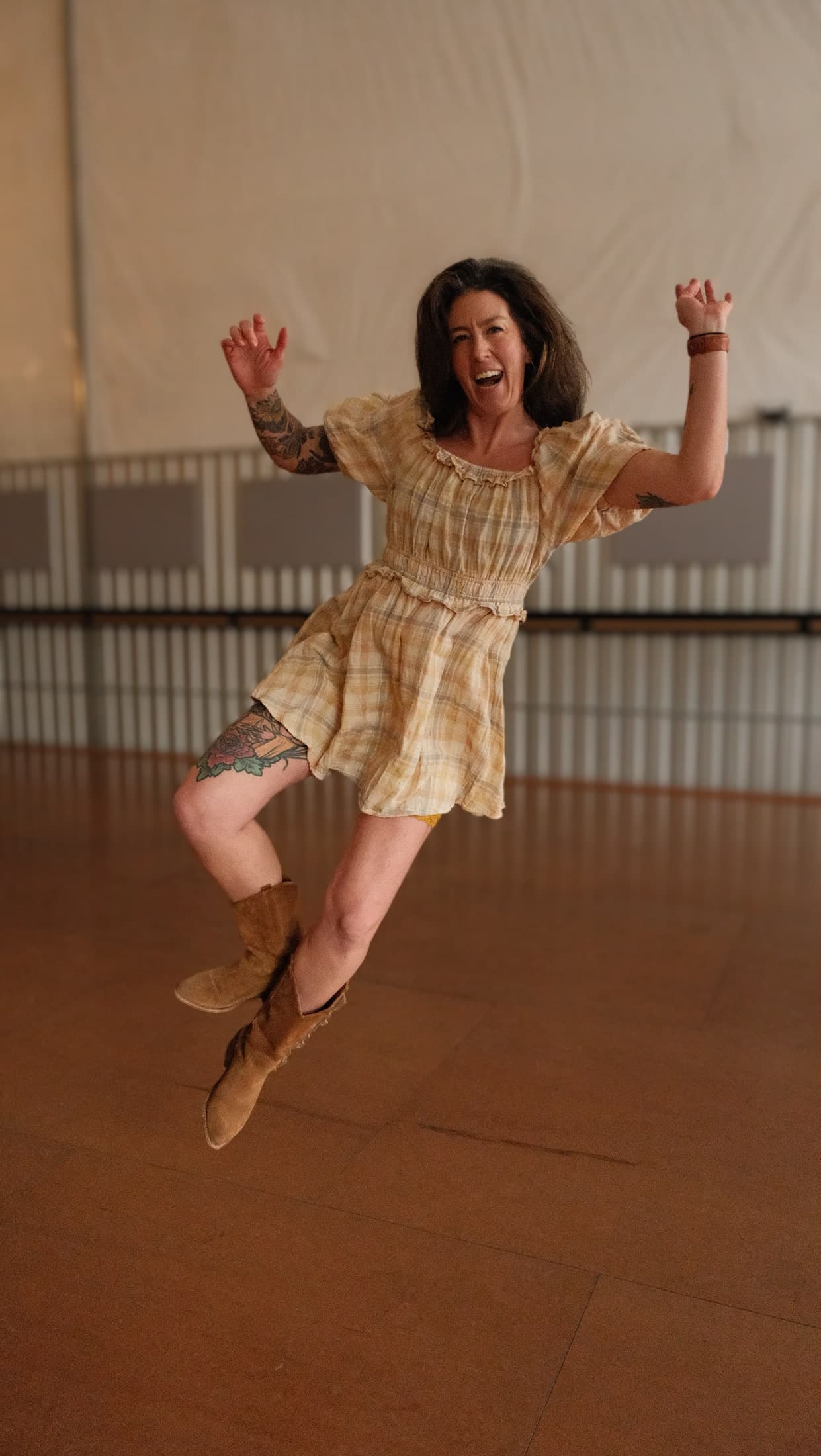 Leigh mid-jump in a plaid dress and cowboy boots, arms up and laughing on a studio dance floor.