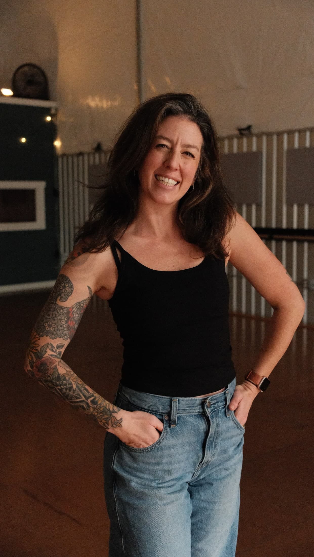 Leigh smiling straight at the camera, hands tucked in her jeans pockets, in a warm dance studio.