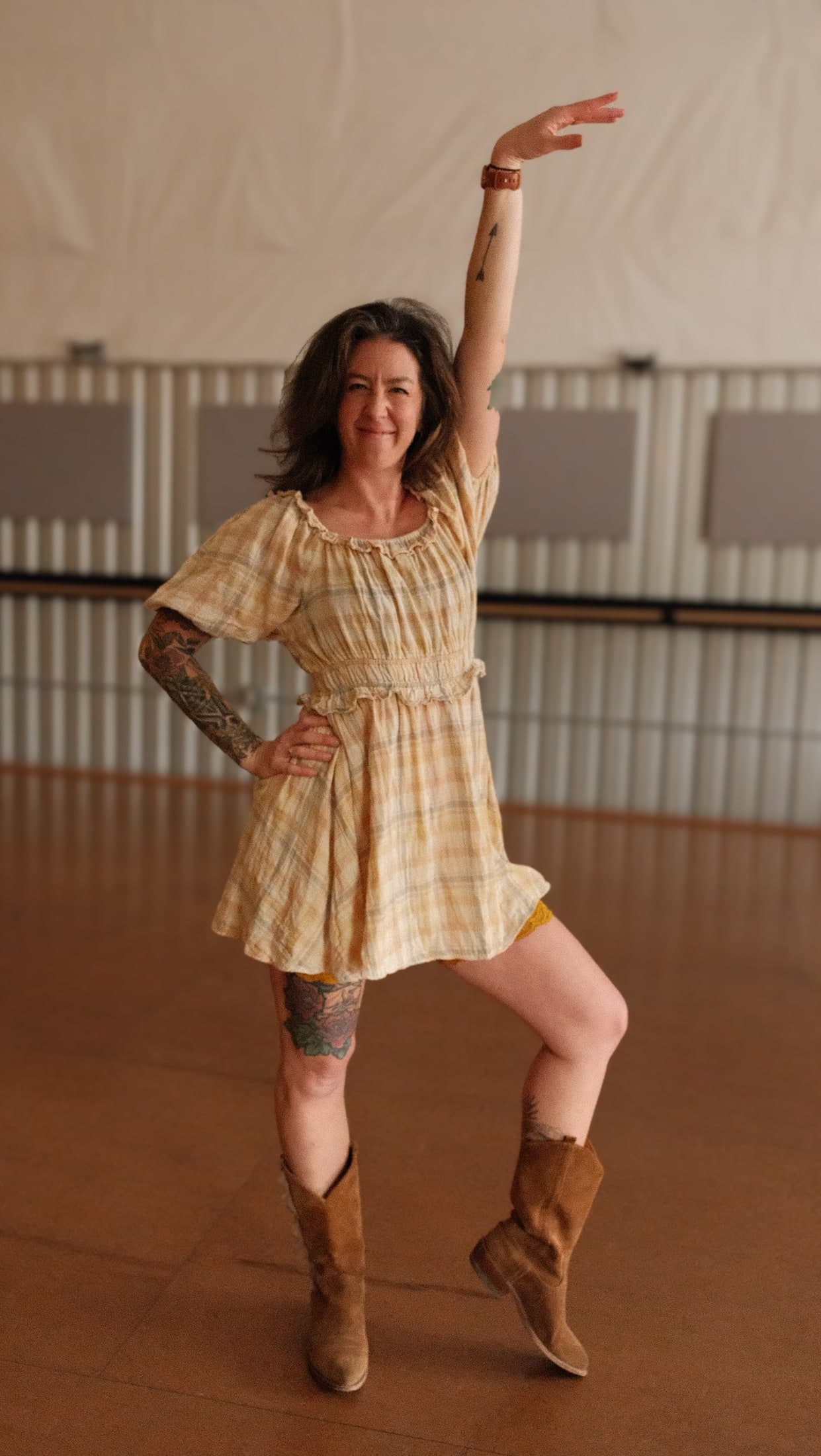 Leigh in a plaid dress and cowboy boots striking a playful dance pose, one arm raised high.