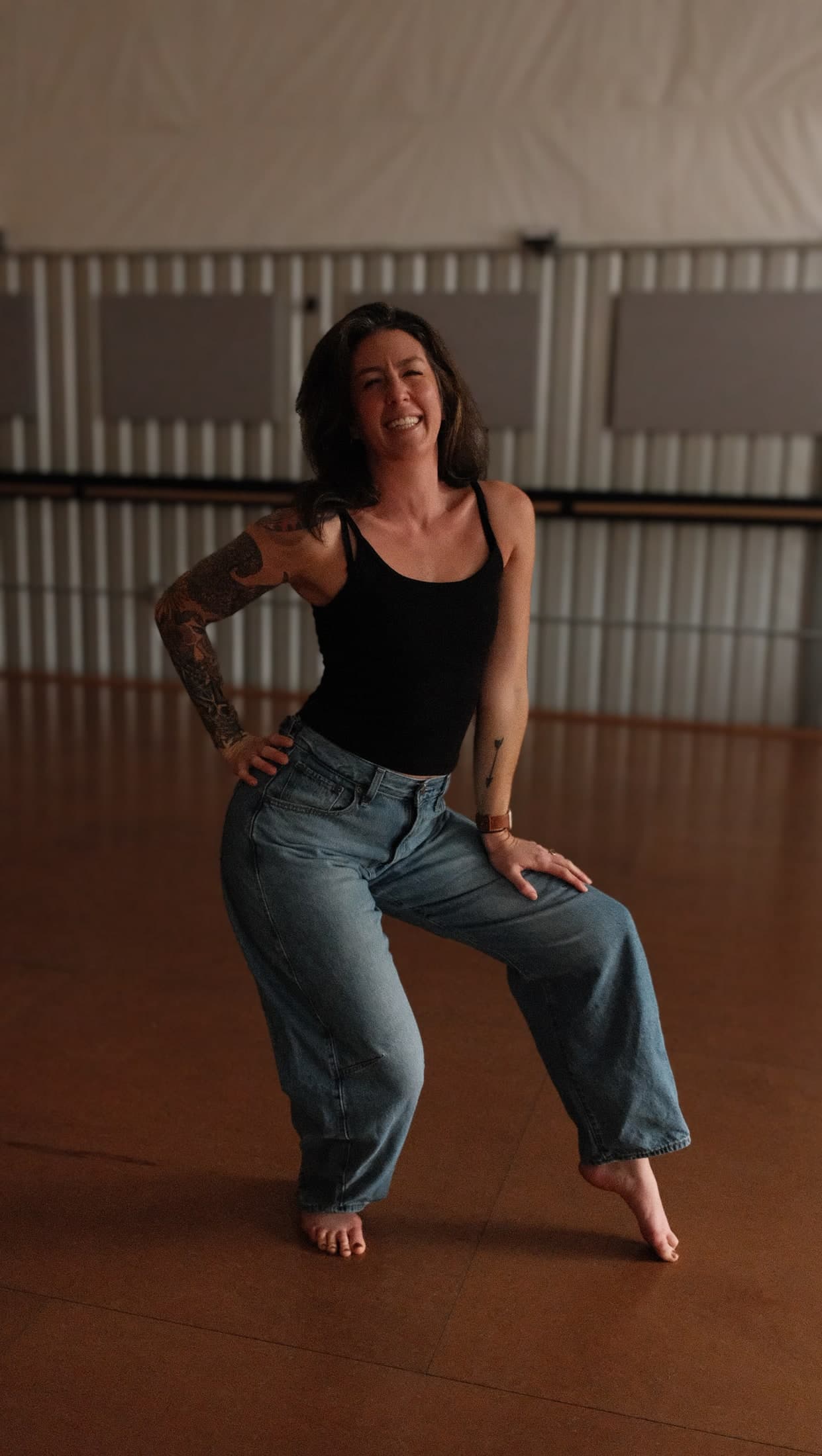 Leigh cracking up mid-dance in a black tank top and jeans, one hand on her hip.